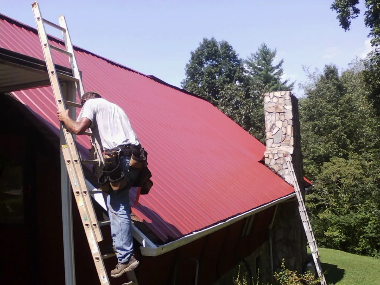 Roofer on a ladder beside a red metal roof and stone chimney — Soffit & Fascia Repair by Roofing Brothers in Lovettsville